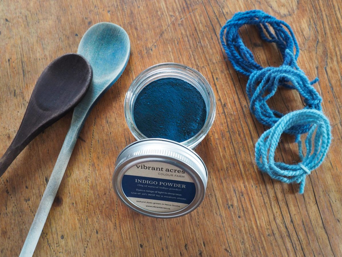 Indigo powder in a jar with Vibrant Acres logo, wooden dye spoons and yarn samples dyed blue